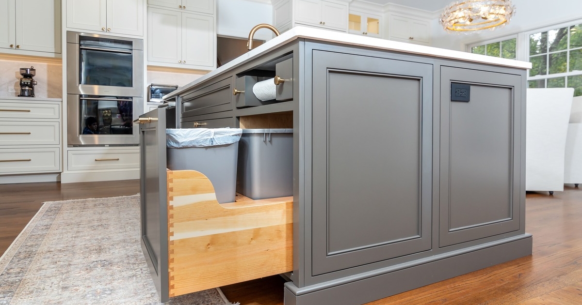 A white kitchen with a island countertop with grey cabinetry and a pullout trash can drawer