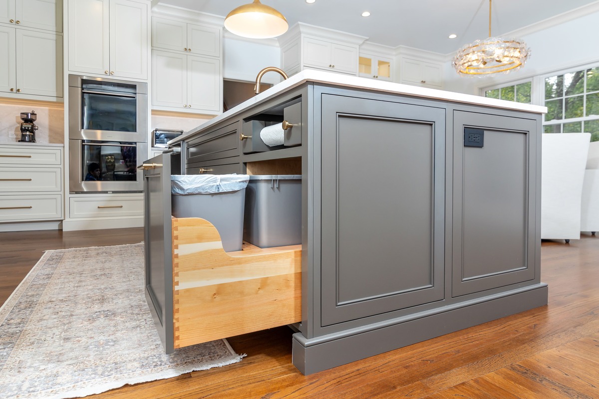 A white kitchen with a island countertop with grey cabinetry and a pullout trash can drawer