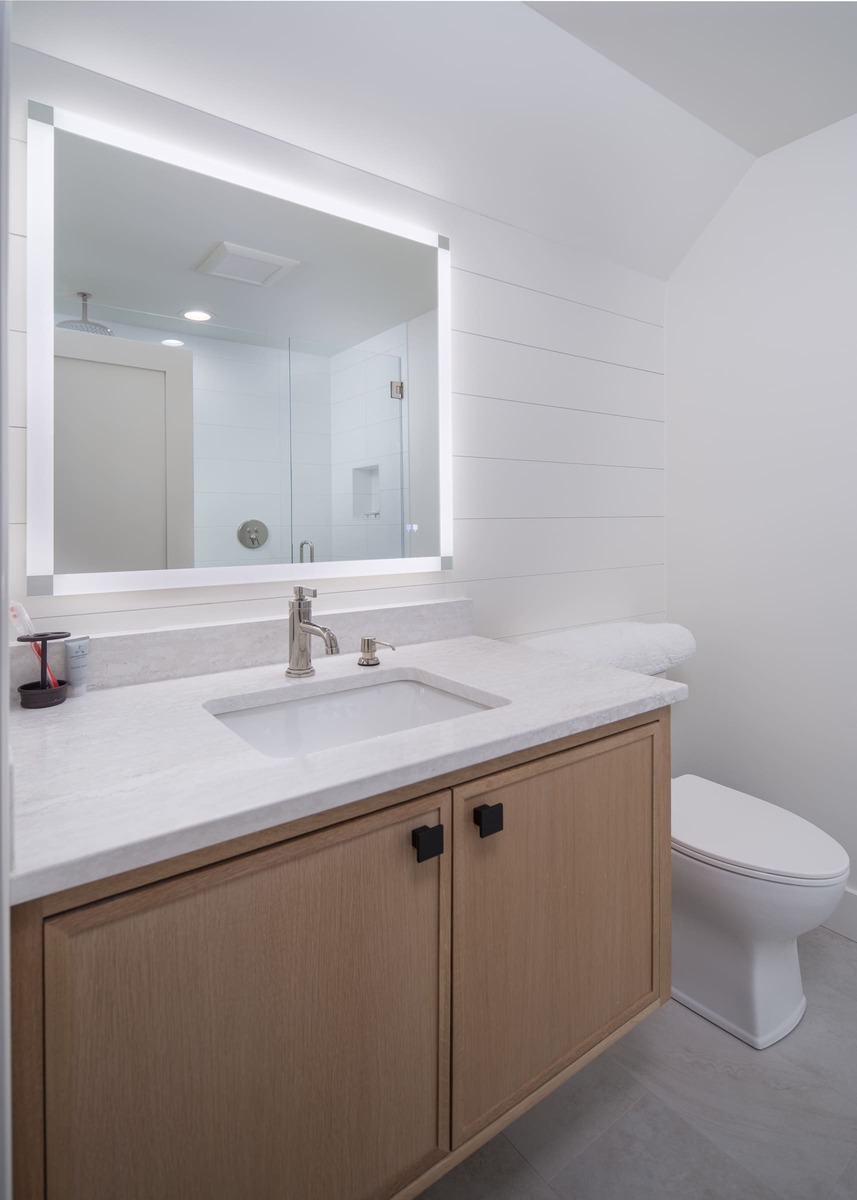 A bathroom with brown cabinets a white countertop with a led mirror and a toilet
