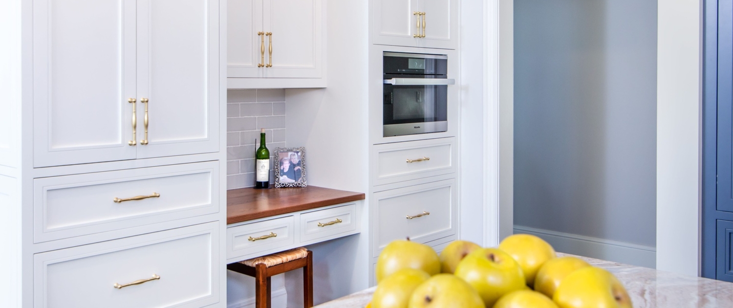A kitchen with white cabinetry and a built in desk and with a steam oven